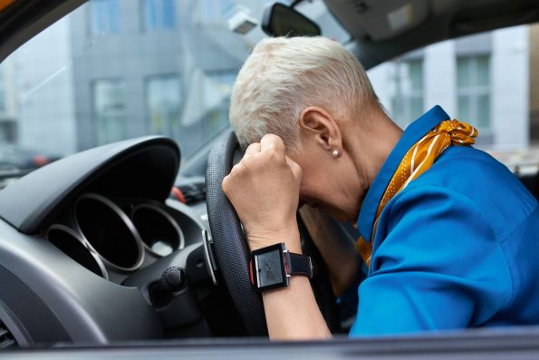 An older woman in a blue outfit with a yellow scarf is sitting in the driver’s seat of a car, resting her head on the steering wheel with her hands clenched in frustration. She appears stressed or fatigued, highlighting the emotional and physical toll of driving.