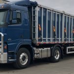 A large blue Volvo semi-truck with a reinforced front grille and an enclosed trailer is parked in an industrial area. The trailer is secured with multiple orange straps and metal bars, designed for transporting bulk materials. The truck features a robust bull bar for added protection, reflecting its use in demanding transport environments. The background includes a warehouse and loading docks, indicating a logistics or freight facility.