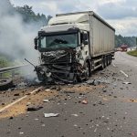 A large white truck with a heavily damaged front end is seen on the side of a highway, surrounded by debris and smoke. A detached wheel and scattered wreckage lie across the road, while emergency vehicles approach in the background. The image highlights the devastating impact of heavy vehicle crashes and the importance of road safety measures.